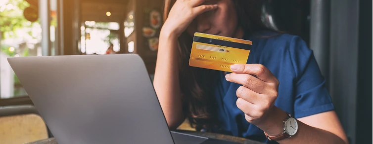 Una mujer muestra frustración frente a su laptop mientras sostiene una tarjeta de crédito, representando el estrés por los retrasos y complicaciones en el recaudo de dinero del exterior.